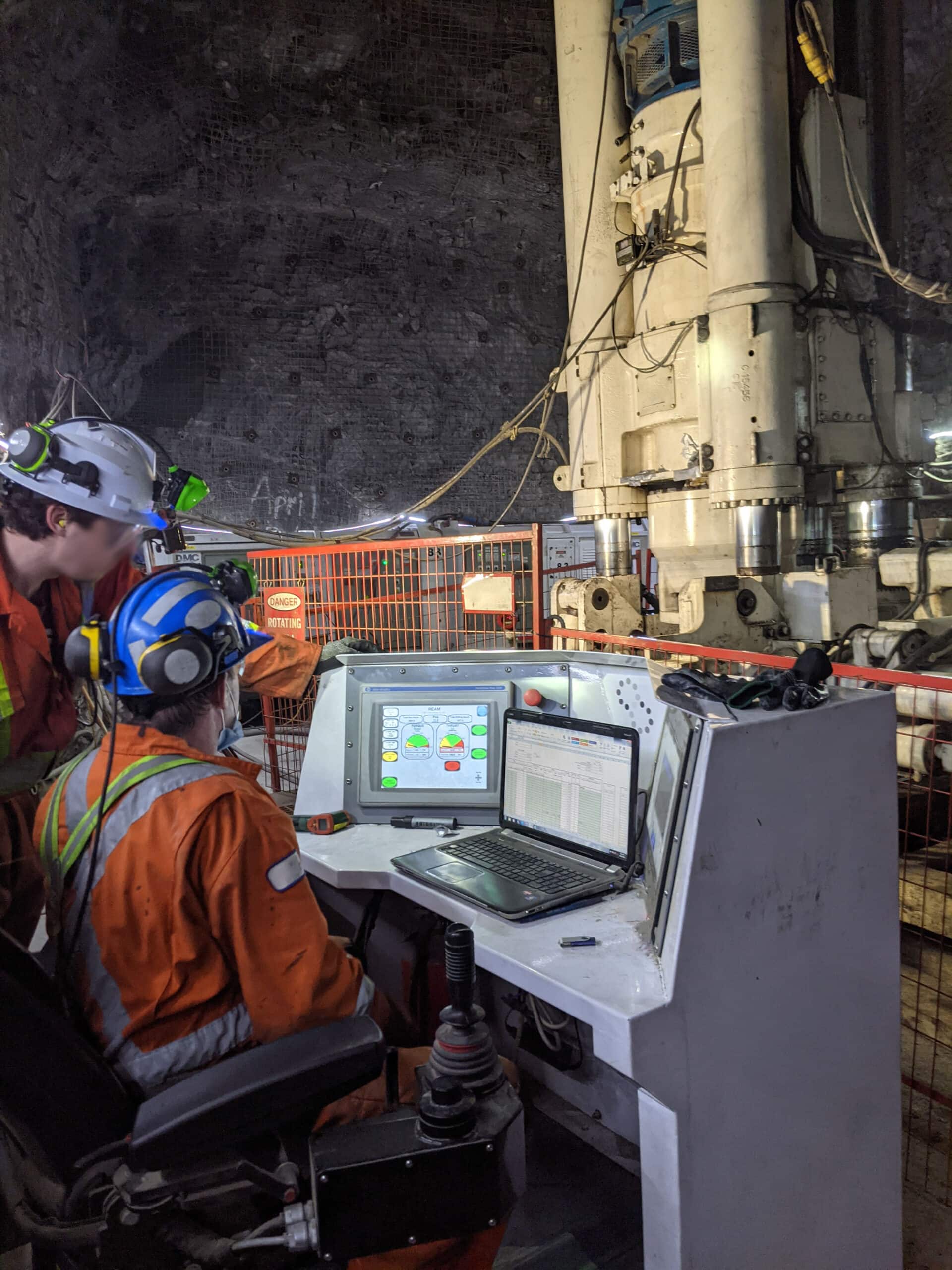 Two workers at a control station overlooping a large drill in an underground mine
