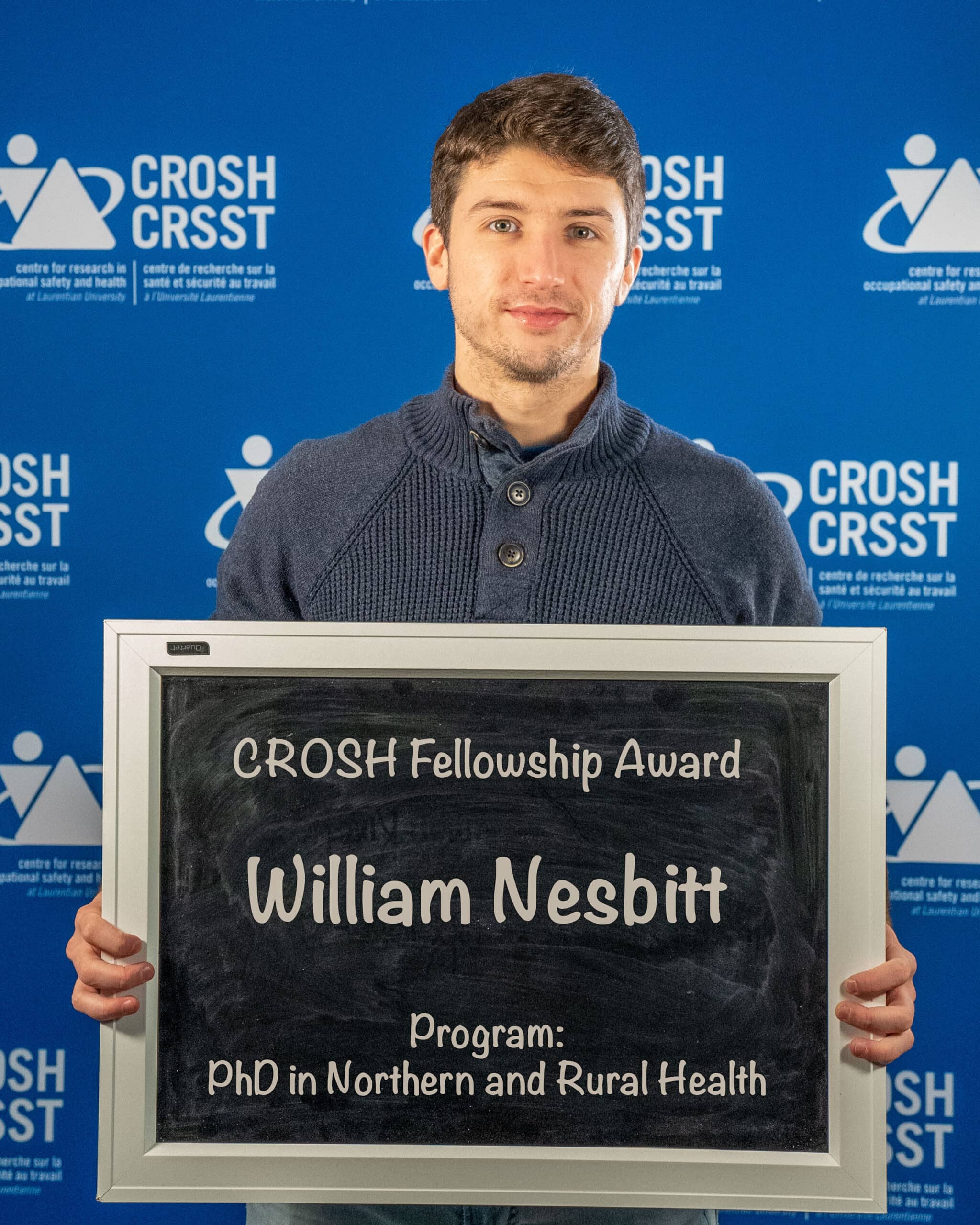 CROSH student member holding a chalkboard that reads "William Nesbitt, CROSH Fellowship Award, Program: PhD in Northern and Rural Health"