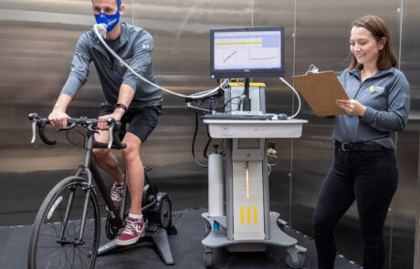 A researcher taking notes on a person riding stationary bike while hooked up to a machine that measures they're breathing