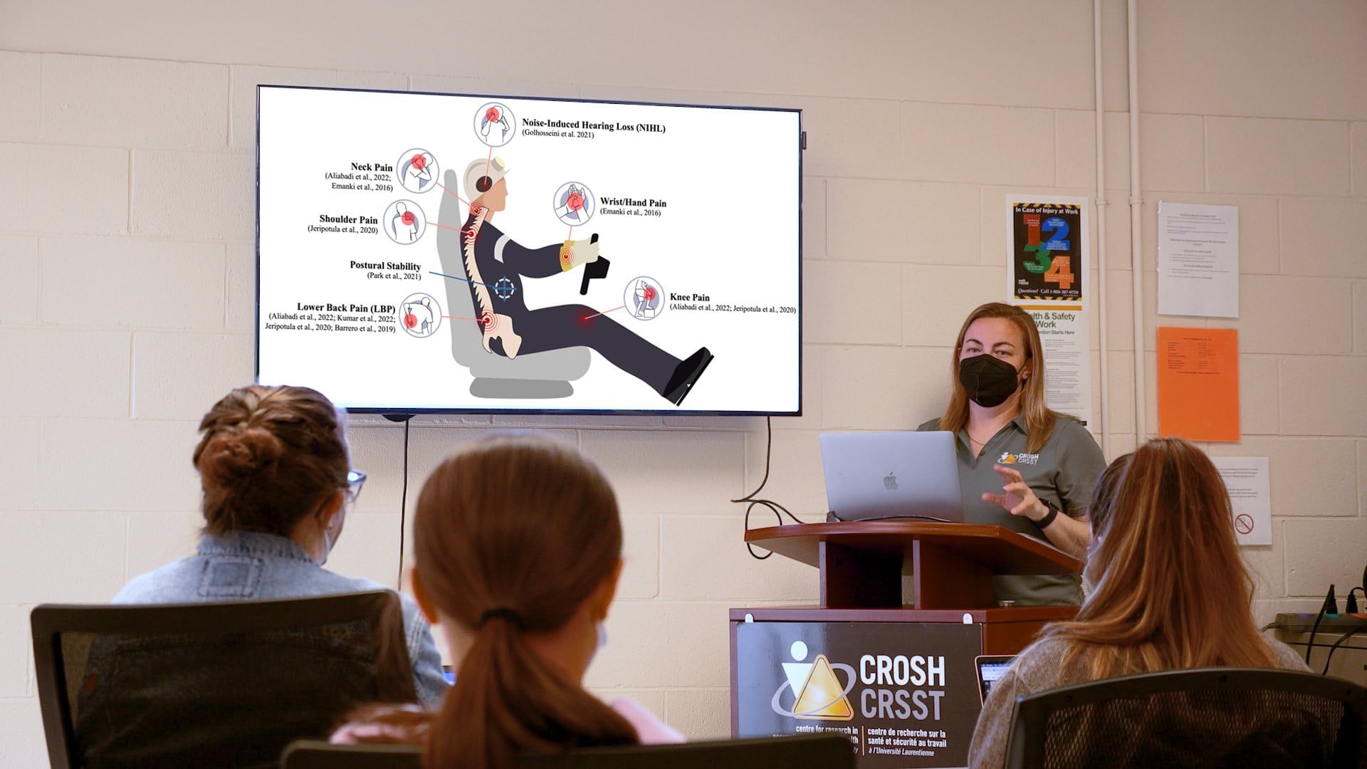 Researcher instructing seated students next to a projected image of the health impacts of vibration exposure.